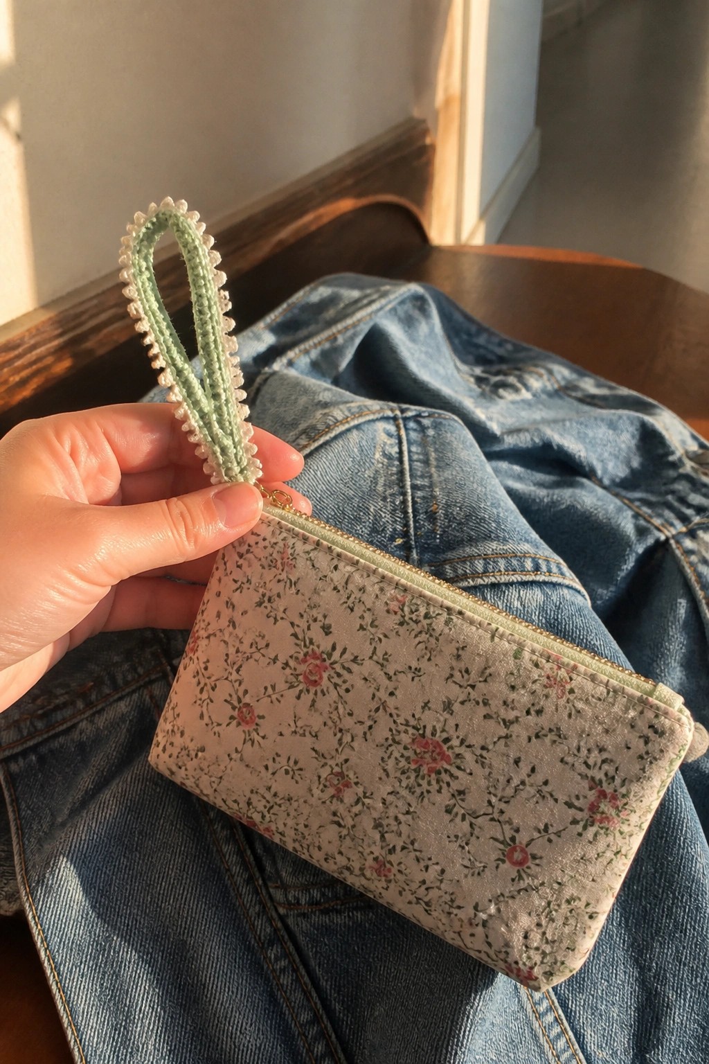 Hand holding floral pouch with crocheted strap atop blue denim jeans