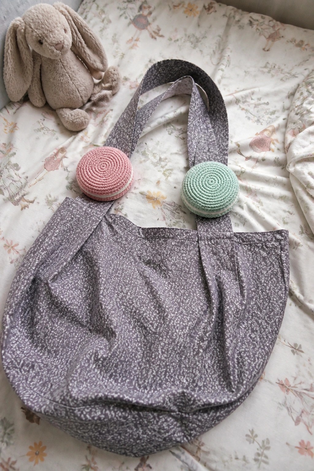 Speckled purple tote bag with crocheted pink and mint macarons beside stuffed bunny on floral bedding.