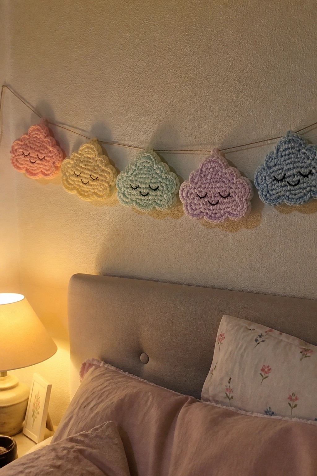 Pastel crocheted cloud garland with faces strung above bed with lamp.
