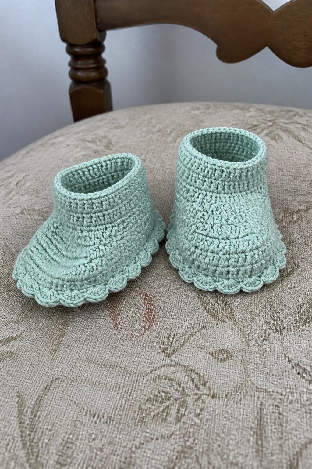 Mint green crocheted baby booties on a beige patterned chair seat