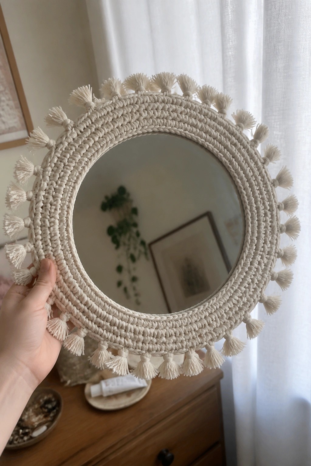 Hand holding round cream macrame mirror with tassels above wooden dresser