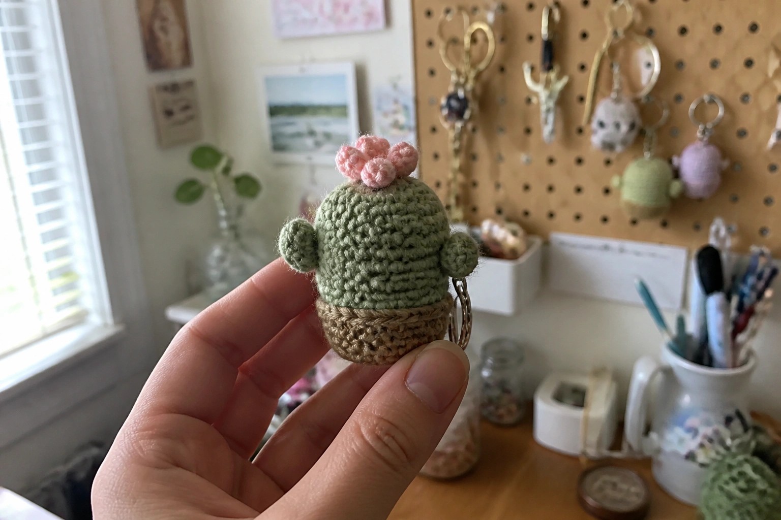 Hand holding crocheted green cactus keychain with pink flowers and brown base.