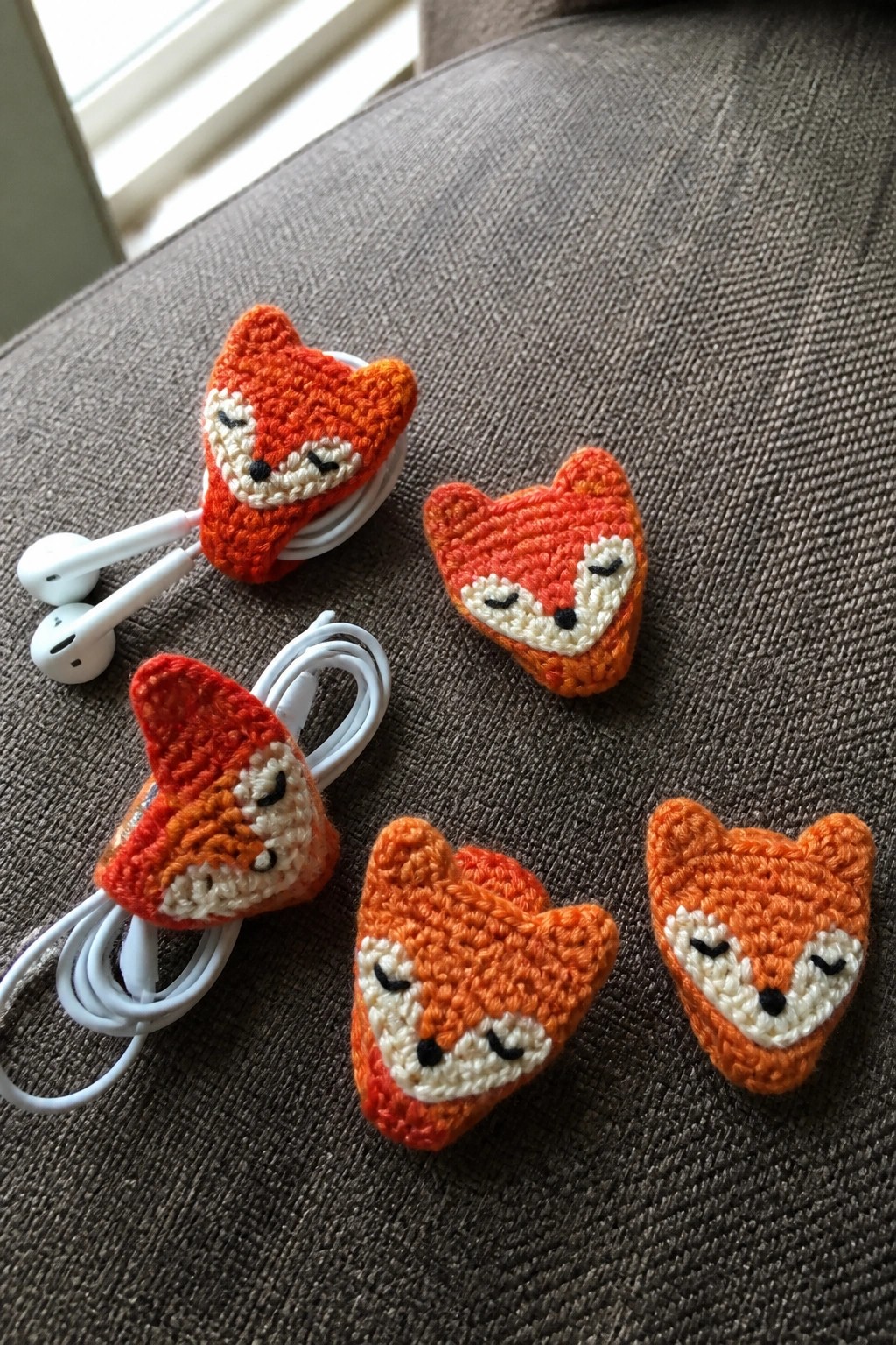 Crocheted orange fox heads holding white earbud cables on brown fabric.