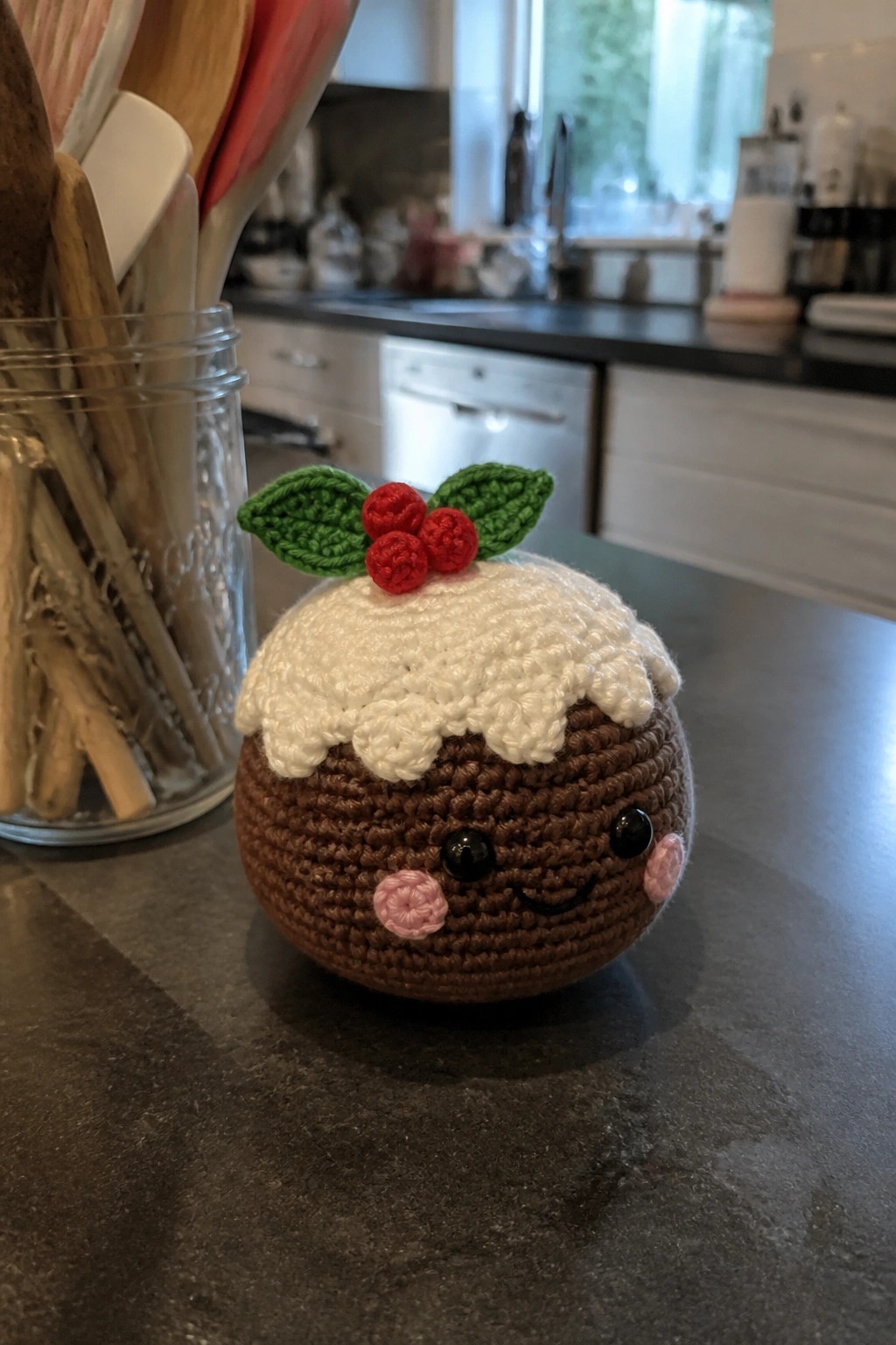 A crocheted Christmas pudding with white top, red berries, green leaves, and a small face rests on a dark countertop.