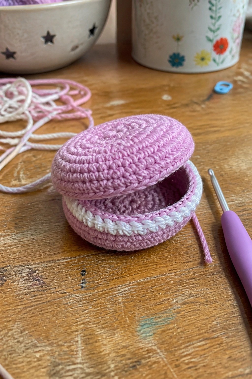 Open pink crocheted pouch with white trim on wooden table beside crochet hook.
