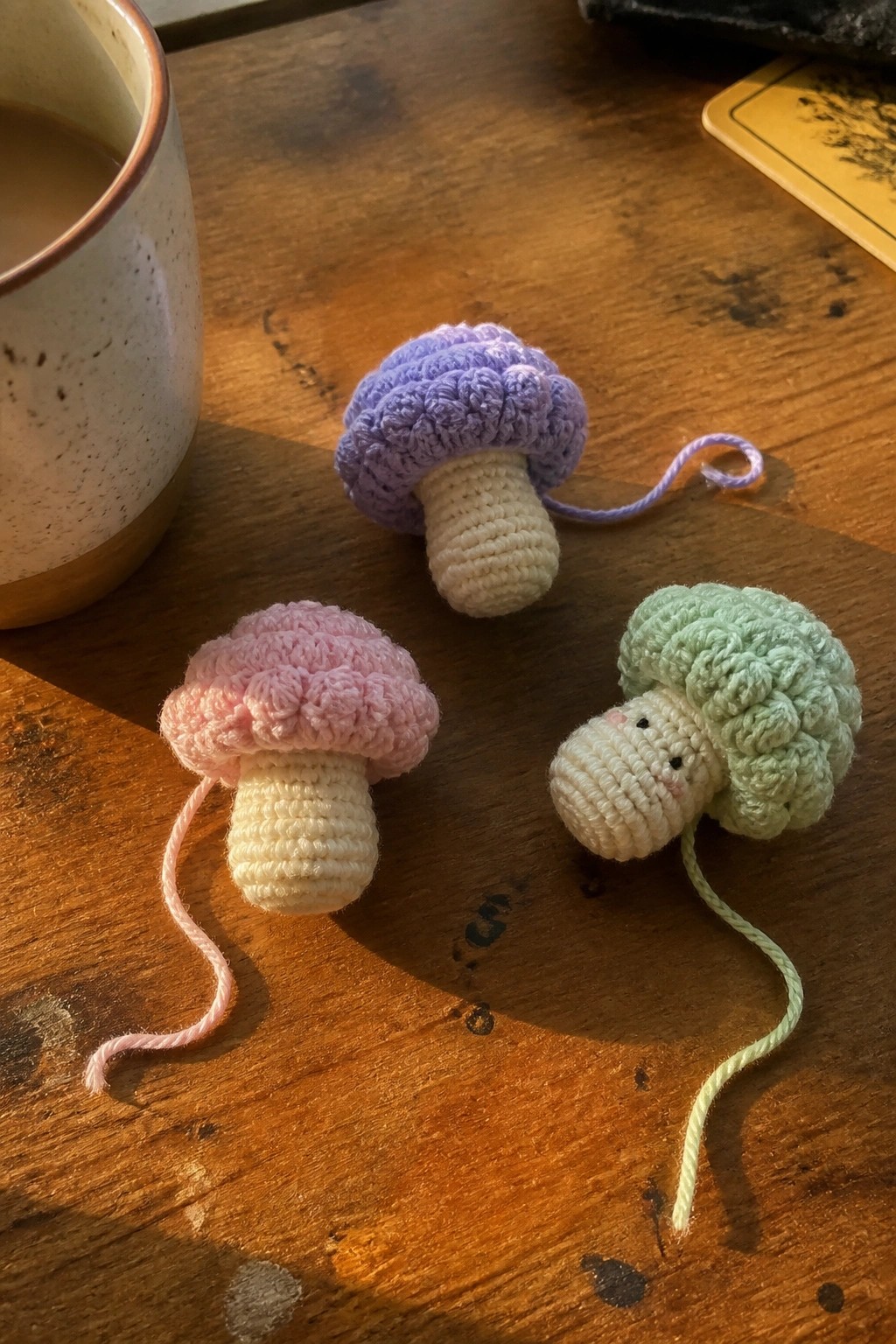 Three small crocheted mushrooms in pink, purple, and mint green with off-white stems rest on a wooden surface beside a ceramic mug.