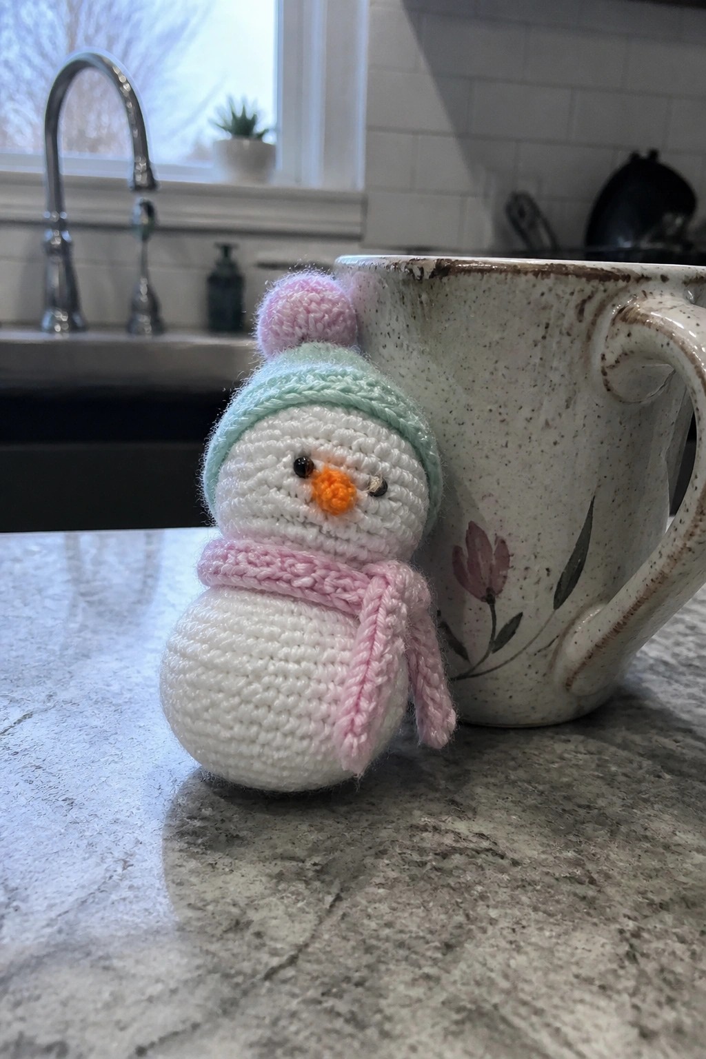 Crocheted snowman with pink scarf and pastel pom-pom hat beside floral mug