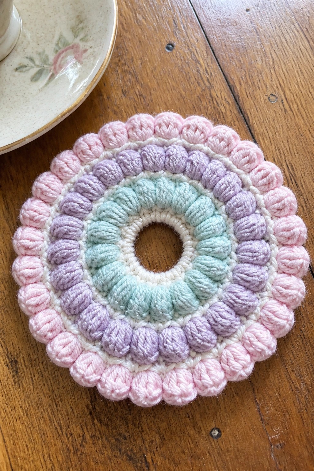 Pastel crocheted circular coaster in pink, purple, mint, and white on wood