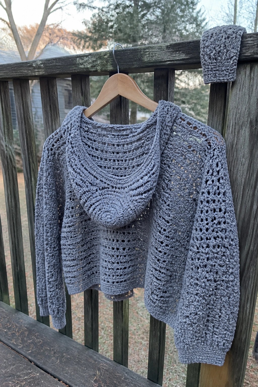 Gray crocheted hooded sweater on wooden hanger over deck railing outdoors
