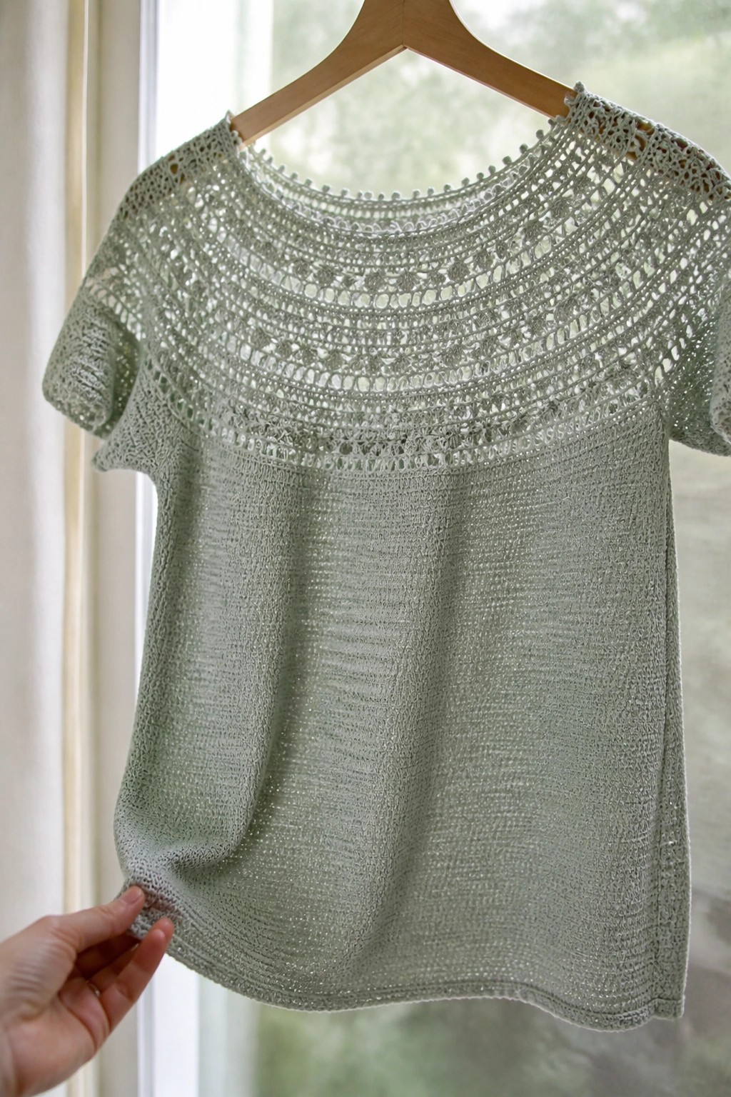 Light green lace-knit top on wooden hanger held near window by hand