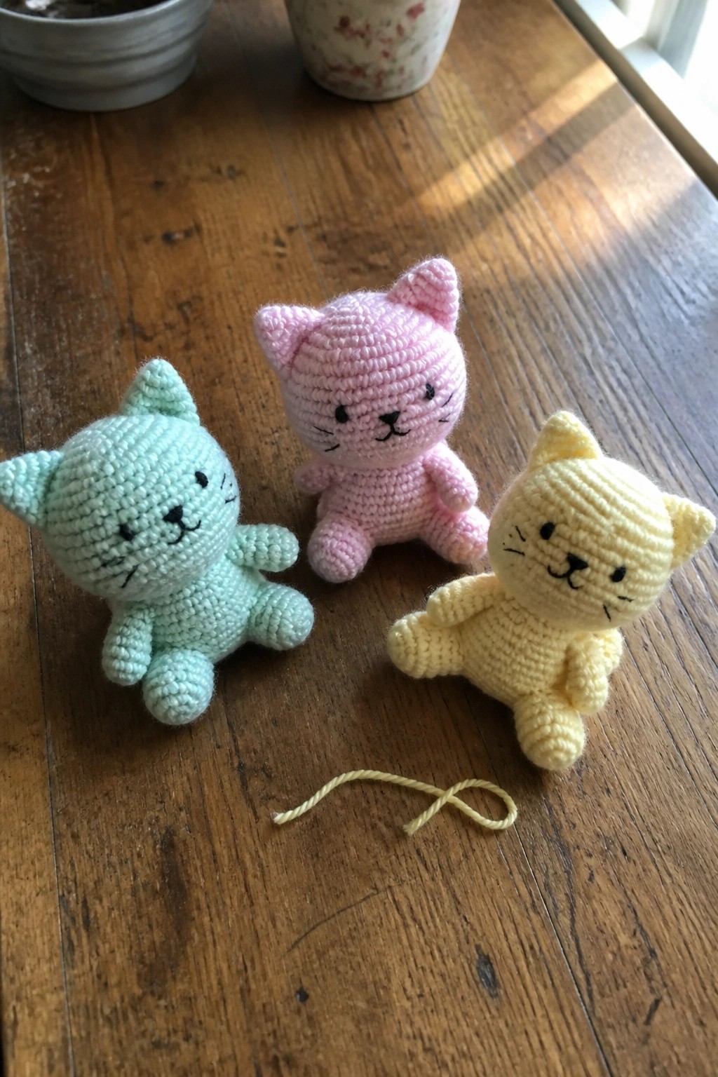 Three pastel crocheted kittens on wooden table with loose yellow yarn nearby