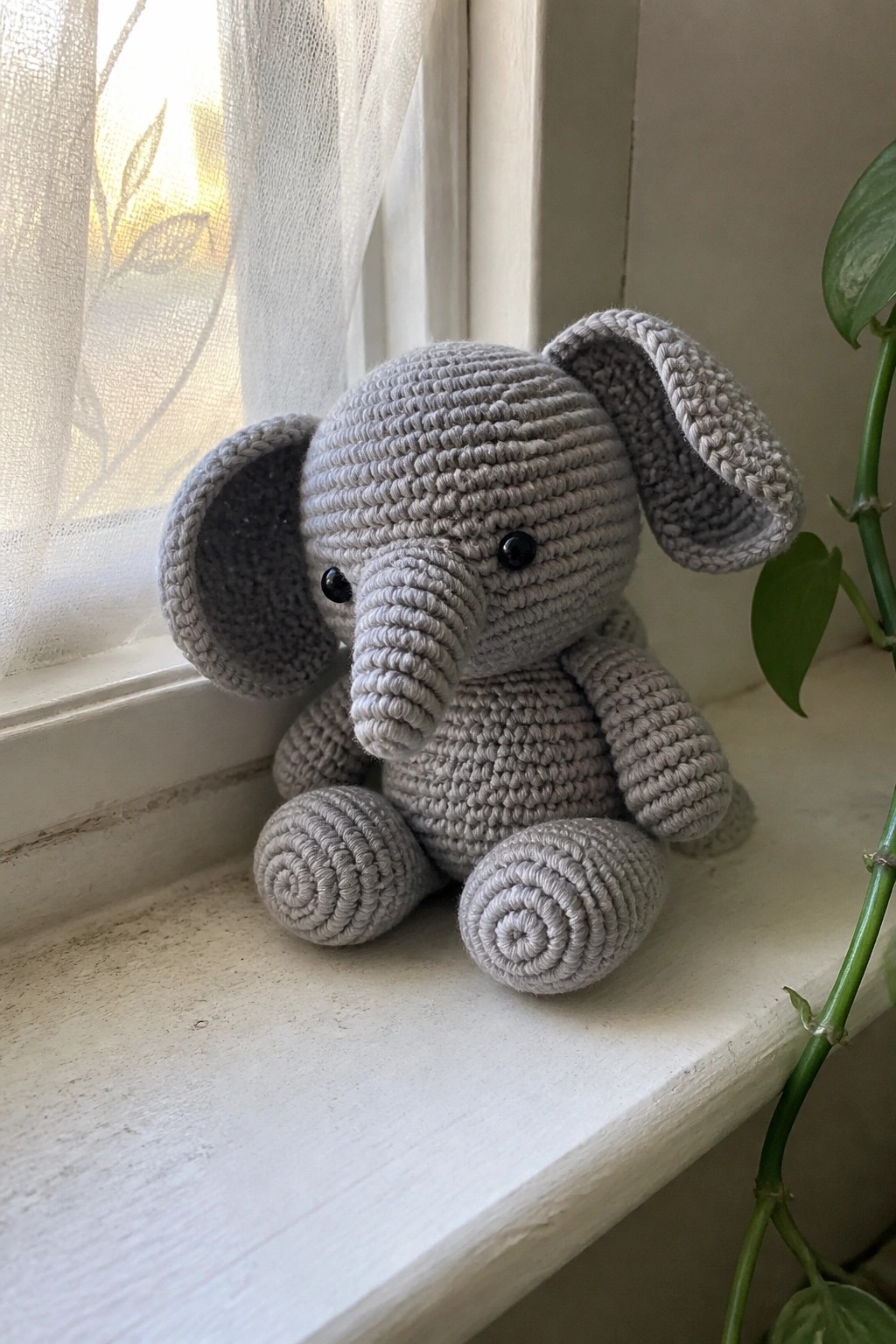 Gray crocheted elephant toy sitting on a sunny windowsill beside a plant.