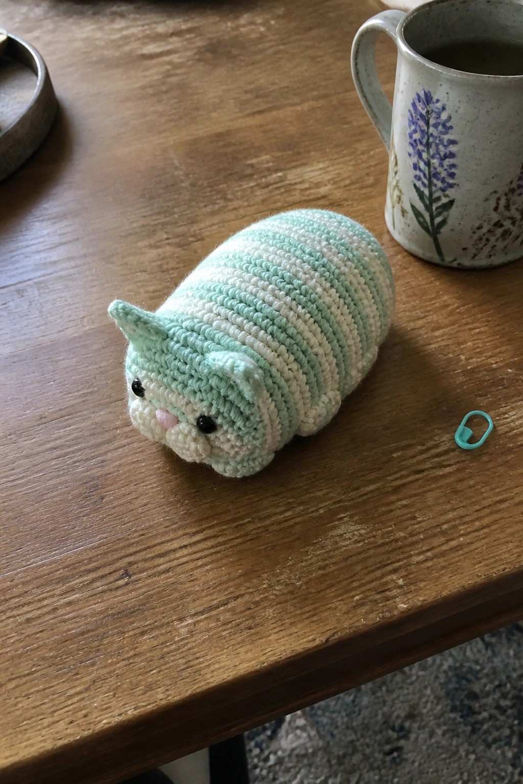 Mint green striped crocheted cat on wooden table beside floral mug