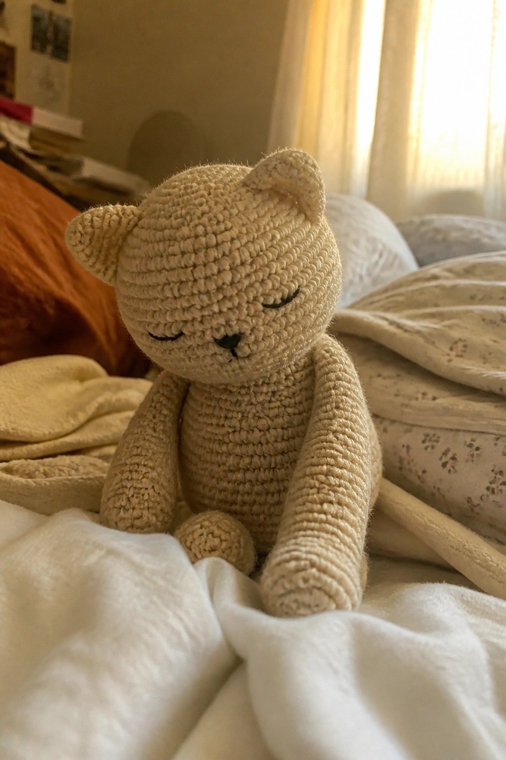 Beige crocheted teddy bear resting on white blankets with sunlight from window