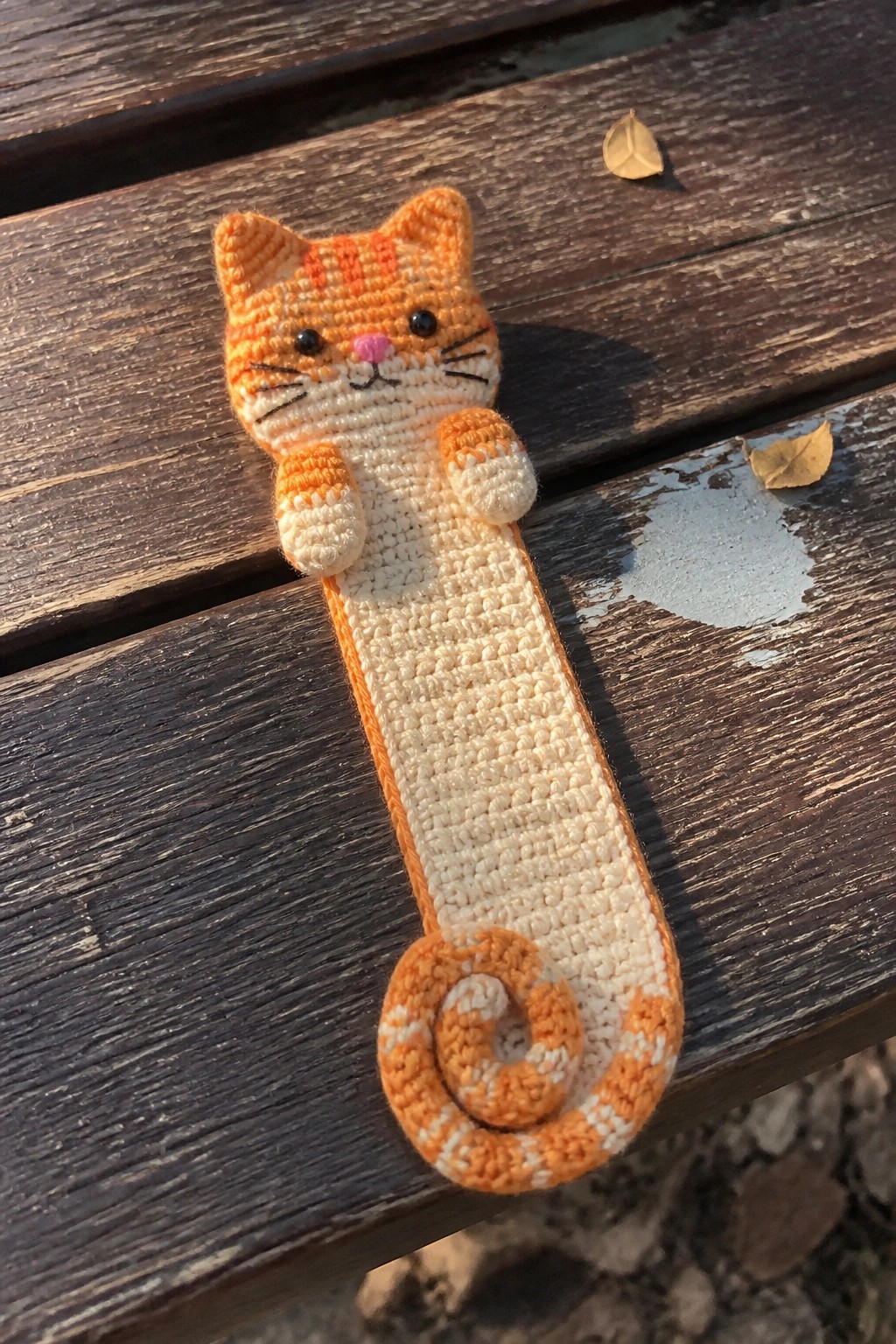 Crocheted orange tabby cat bookmark with raised paws and curled tail on wood.