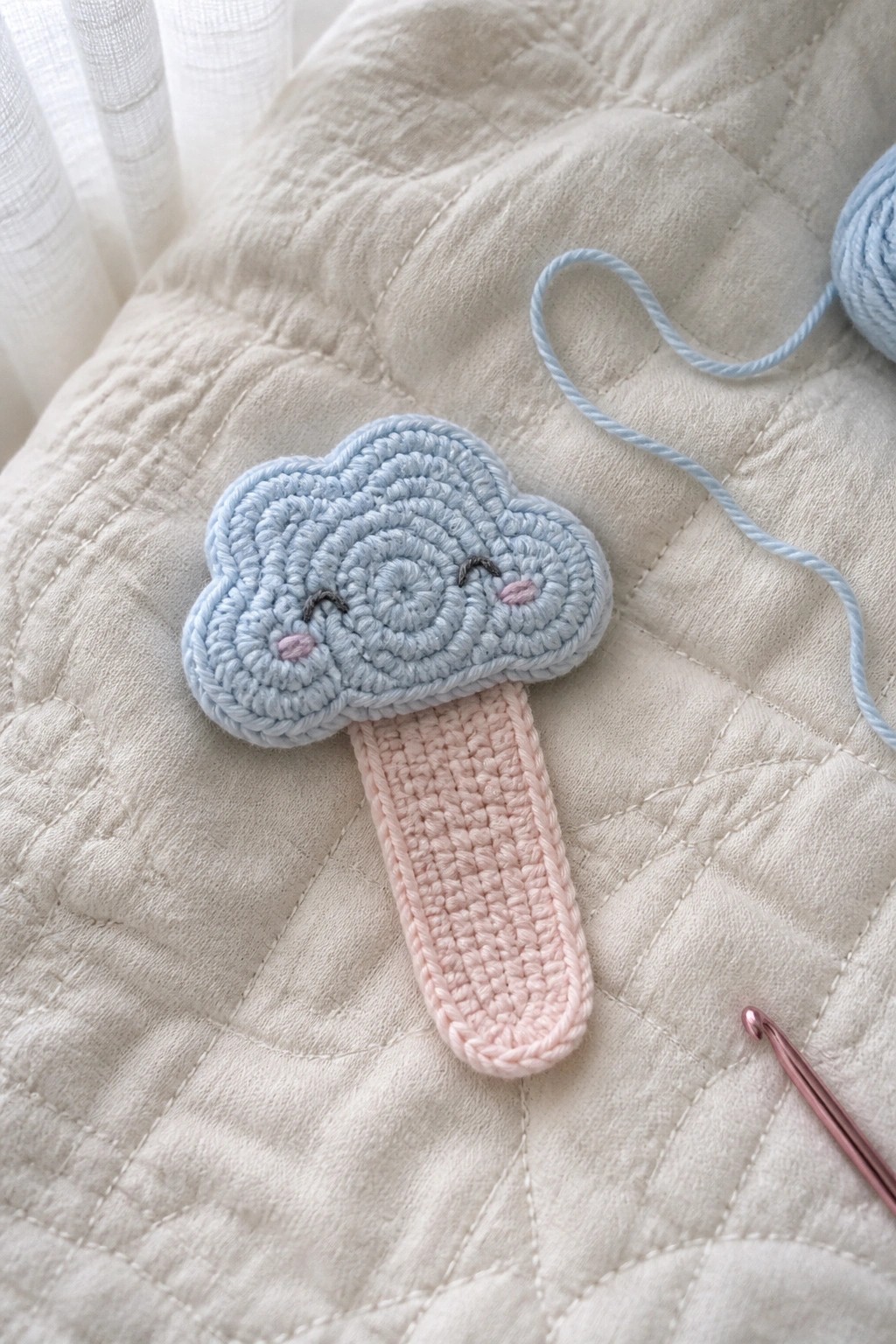 Crocheted light blue cloud with pink handle on white quilted fabric beside hook