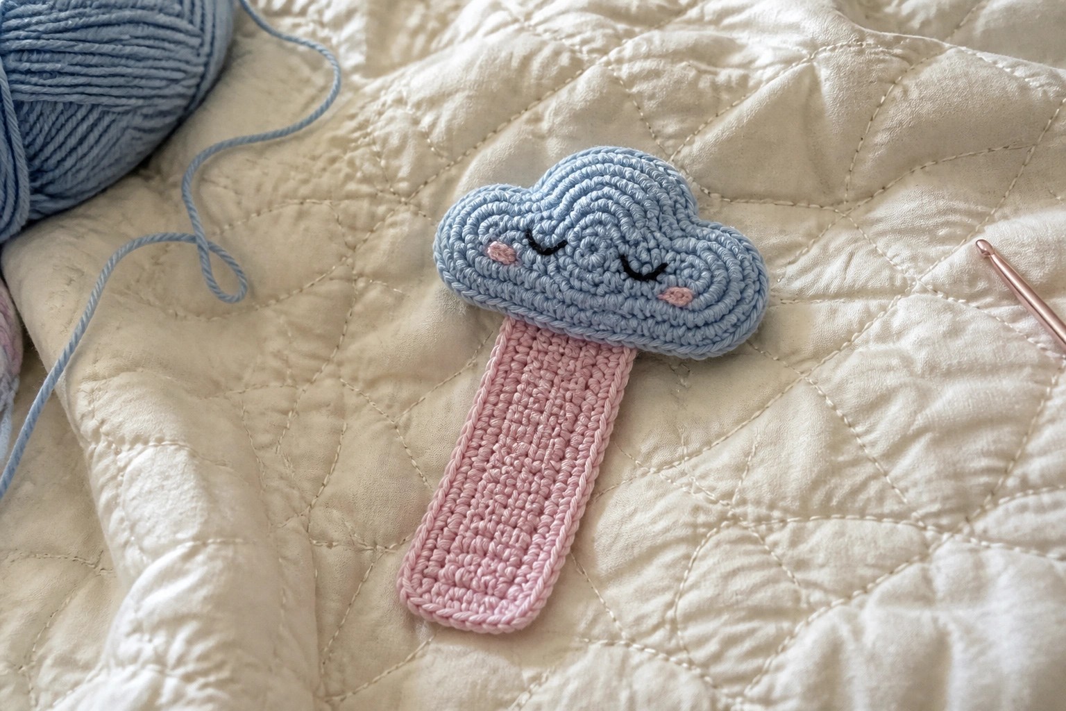 Crocheted light blue cloud with pink handle on white quilted fabric beside hook