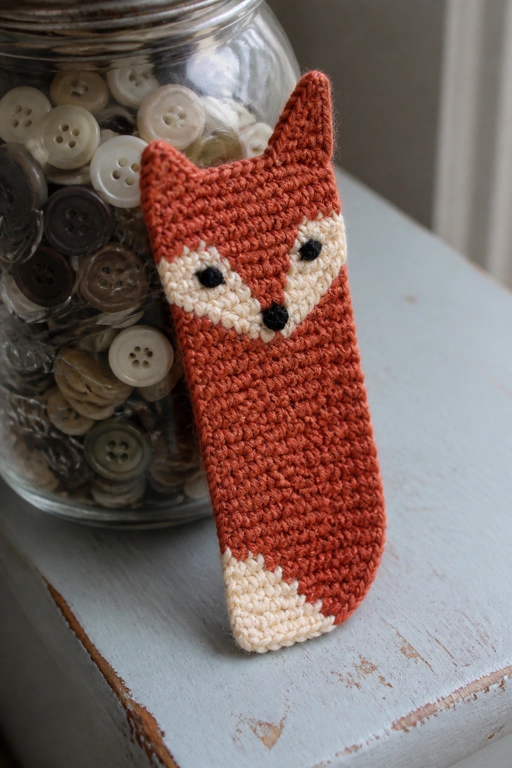Crocheted orange fox beside glass jar of buttons on blue wooden surface