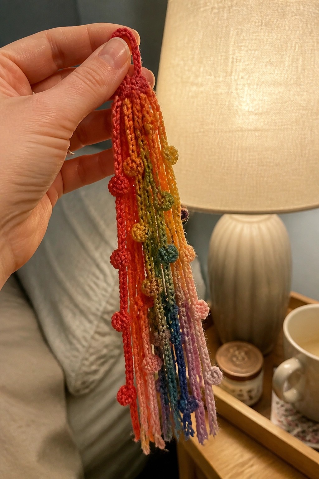 Hand holding rainbow crocheted tassel with colorful pom-poms and fringe strands.