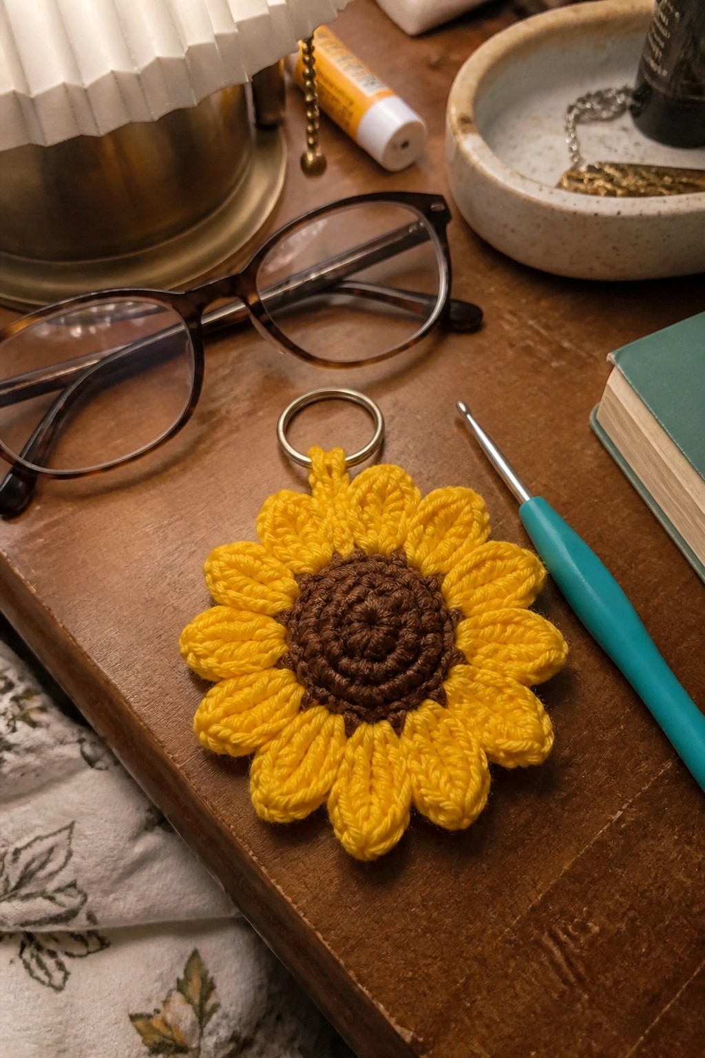 Crocheted yellow sunflower keychain with brown center on wooden table near glasses and hook