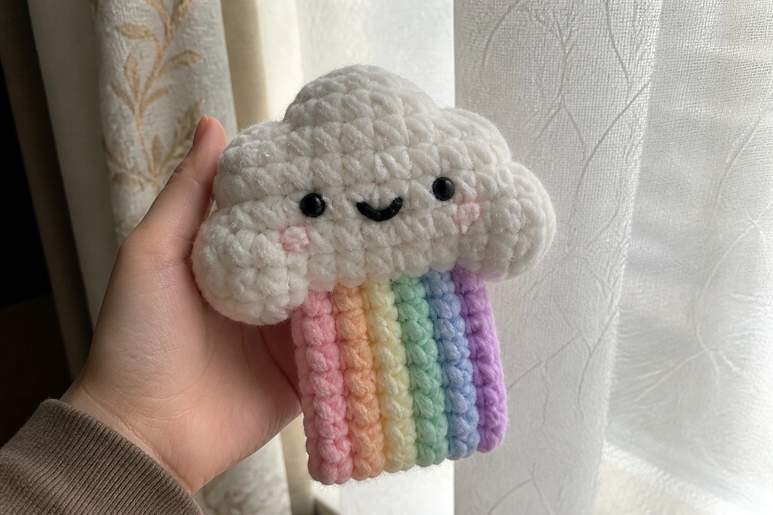 Hand holding white crocheted cloud plush with rainbow yarn strands and smiling face.