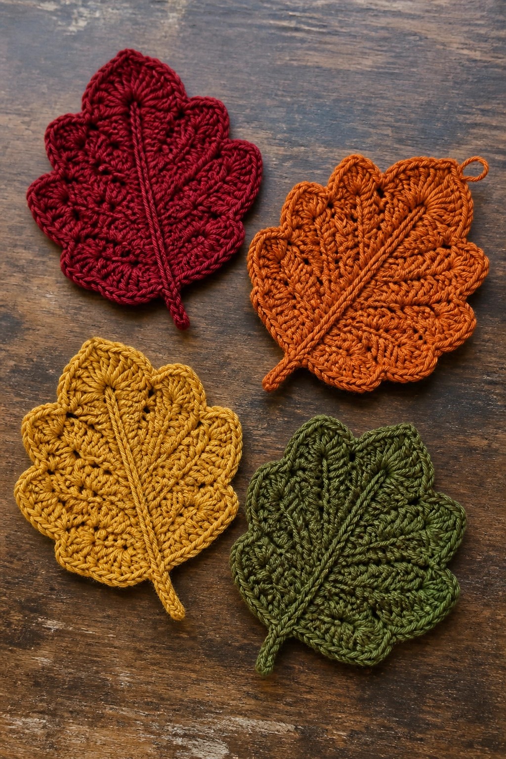 Four crocheted autumn leaves in red, orange, yellow, and green on wood.