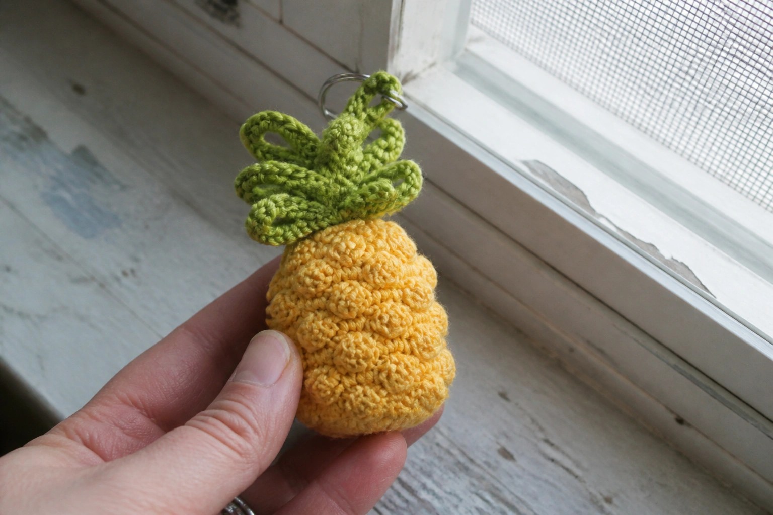 Hand holding yellow crocheted pineapple keychain with green top by windowsill.