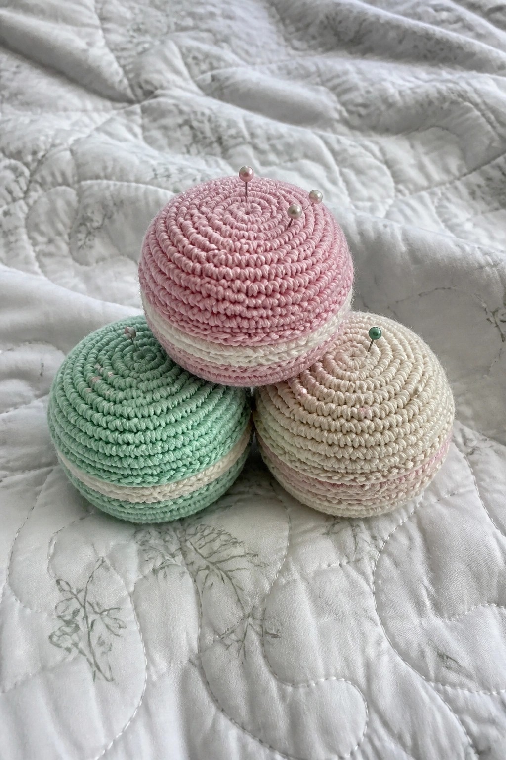 Three pastel crocheted pincushions with pins on a white quilted fabric.