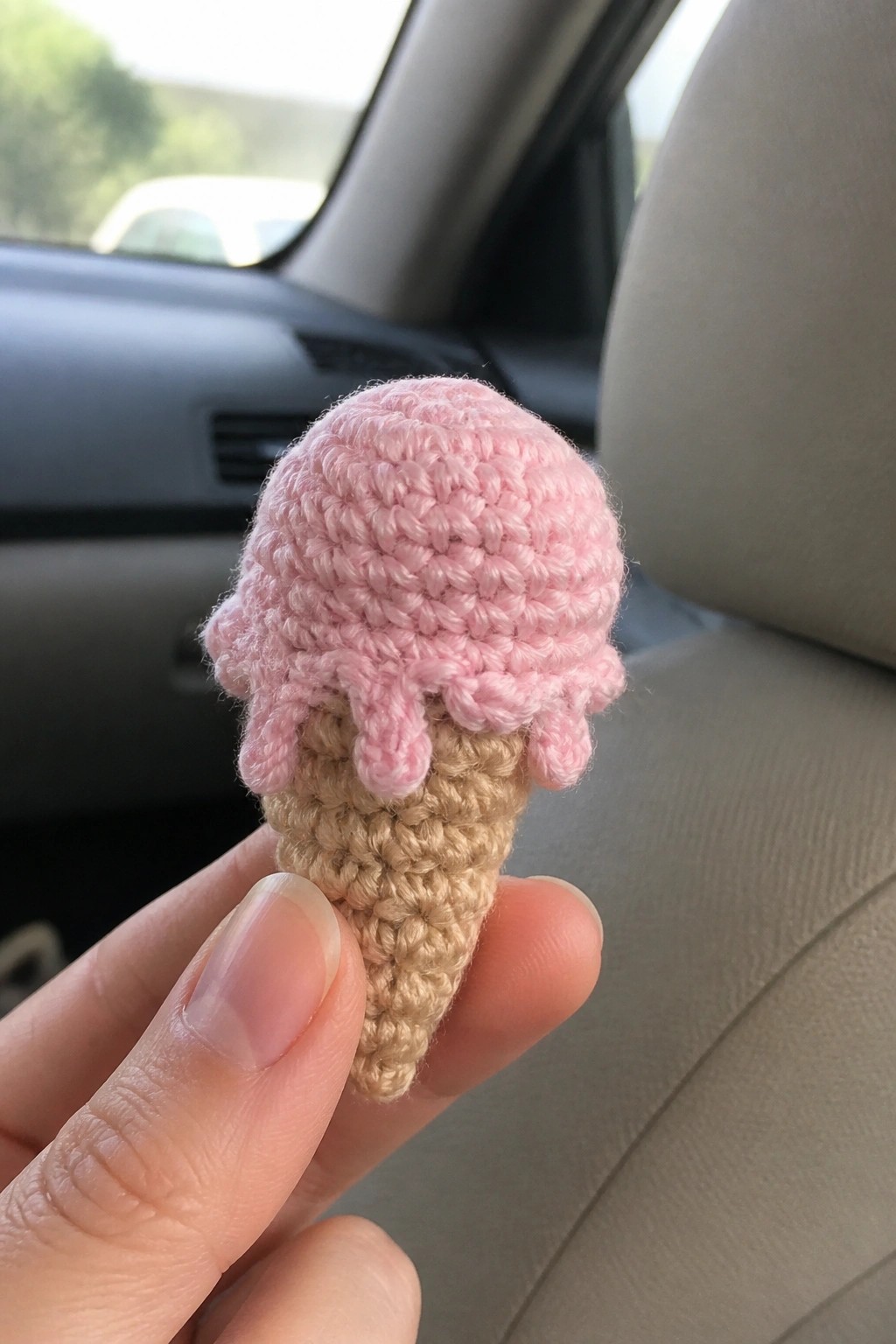 Hand holding crocheted pink melting ice cream on beige cone inside car