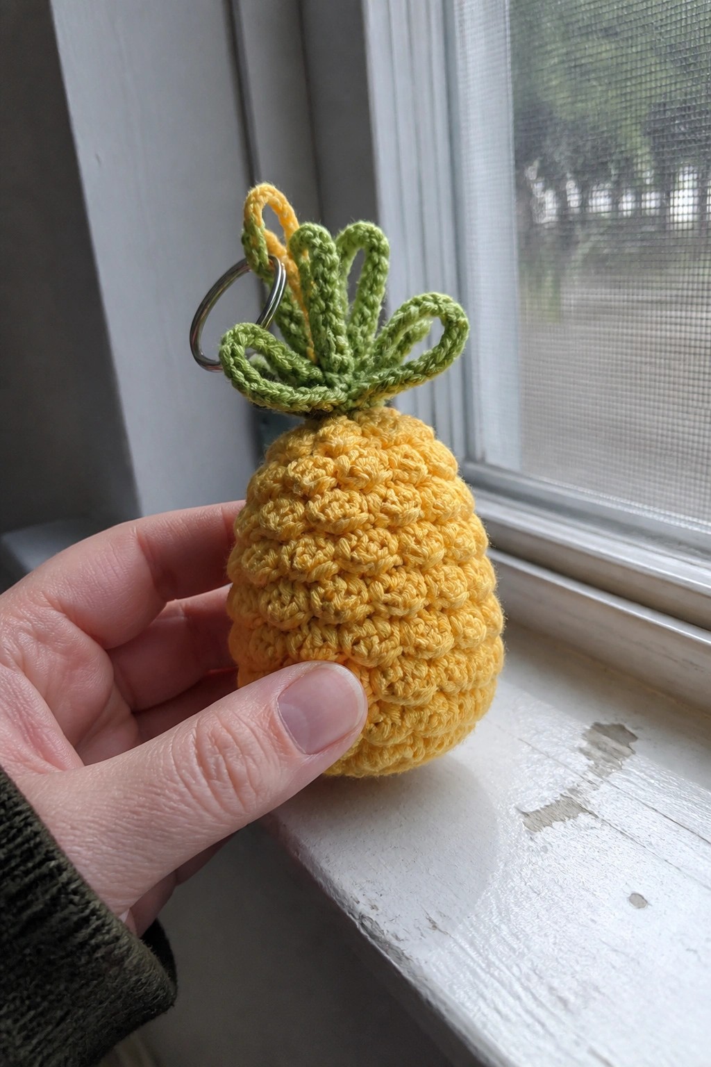 Hand holding yellow crocheted pineapple keychain with green top by windowsill.