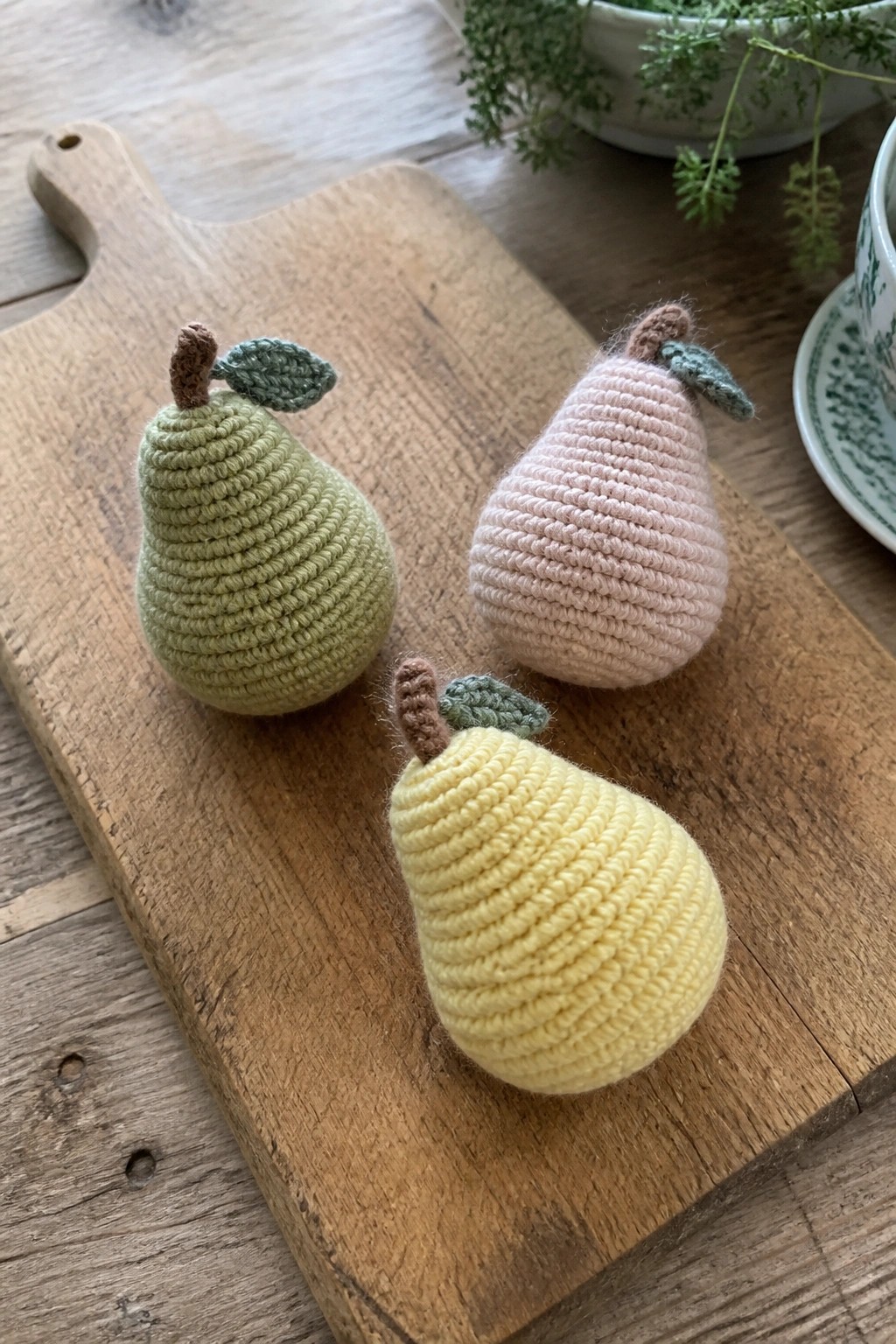 Three crocheted pears in green, pink, and yellow on a wooden cutting board.