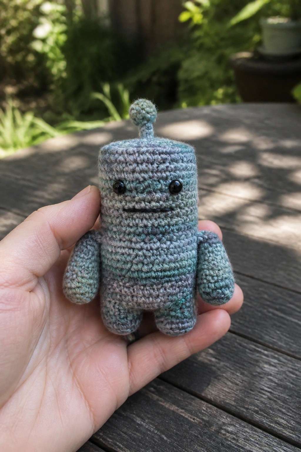 Hand holding small crocheted robot toy in blue-green variegated yarn outdoors.