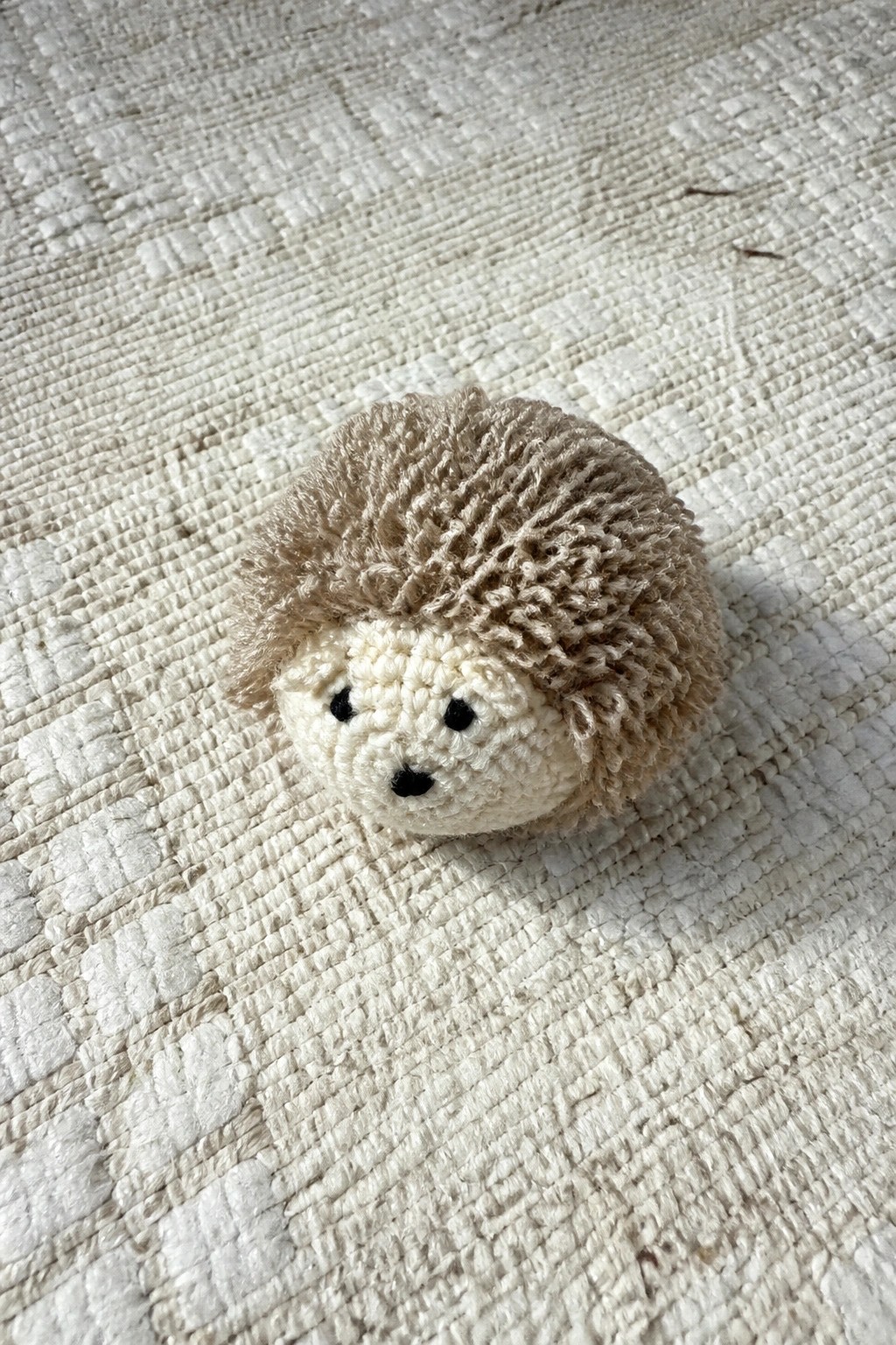 Crocheted beige hedgehog with black eyes and nose on white textured fabric