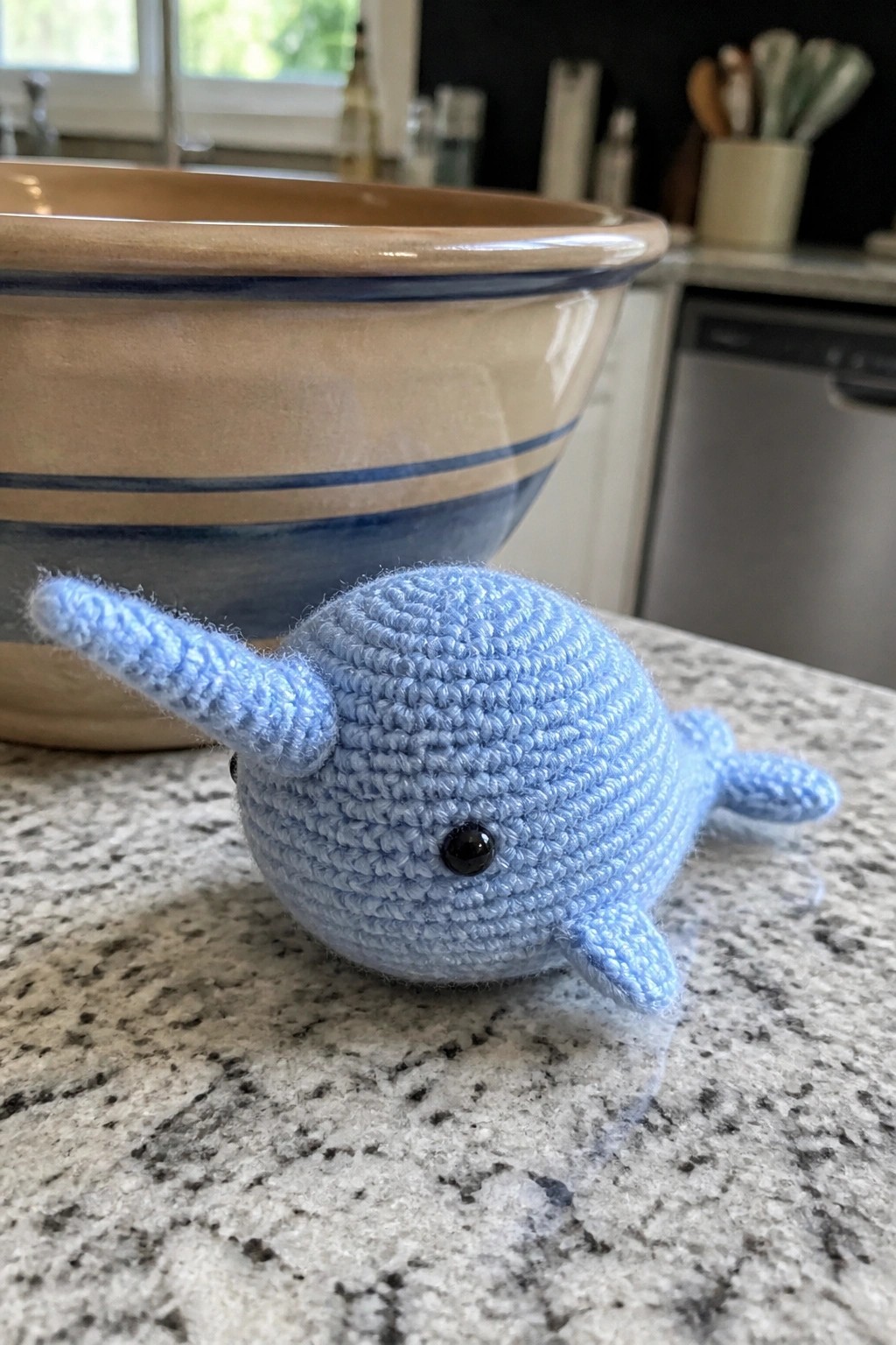 Light blue crocheted narwhal toy on granite countertop near striped bowl.
