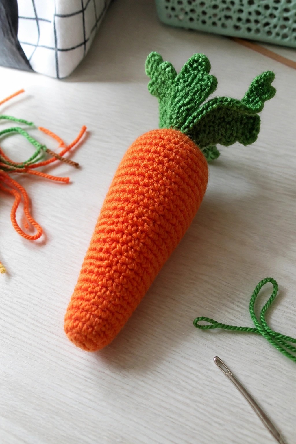 Crocheted orange carrot with green top beside needle and yarn on wooden table