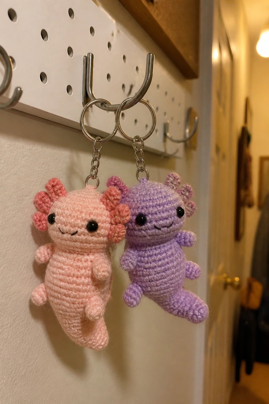 Pink and purple crocheted axolotl keychains hanging from metal rings on pegboard hook