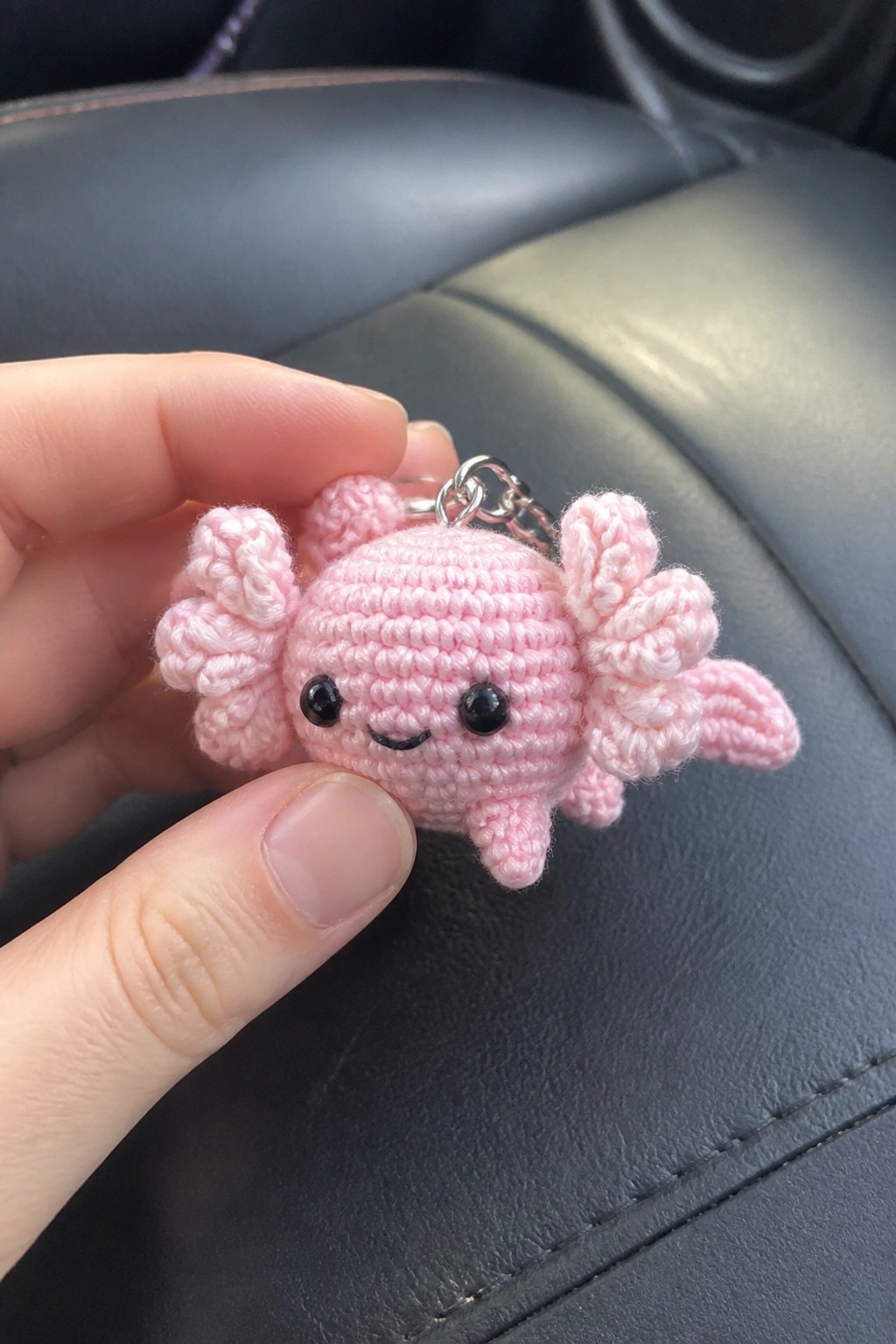 Hand holding pink crocheted axolotl keychain with black eyes on car seat.