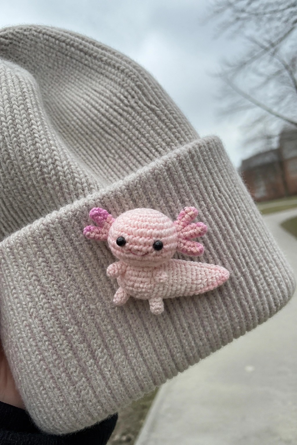 Pink crocheted axolotl attached to cuff of beige knitted beanie.