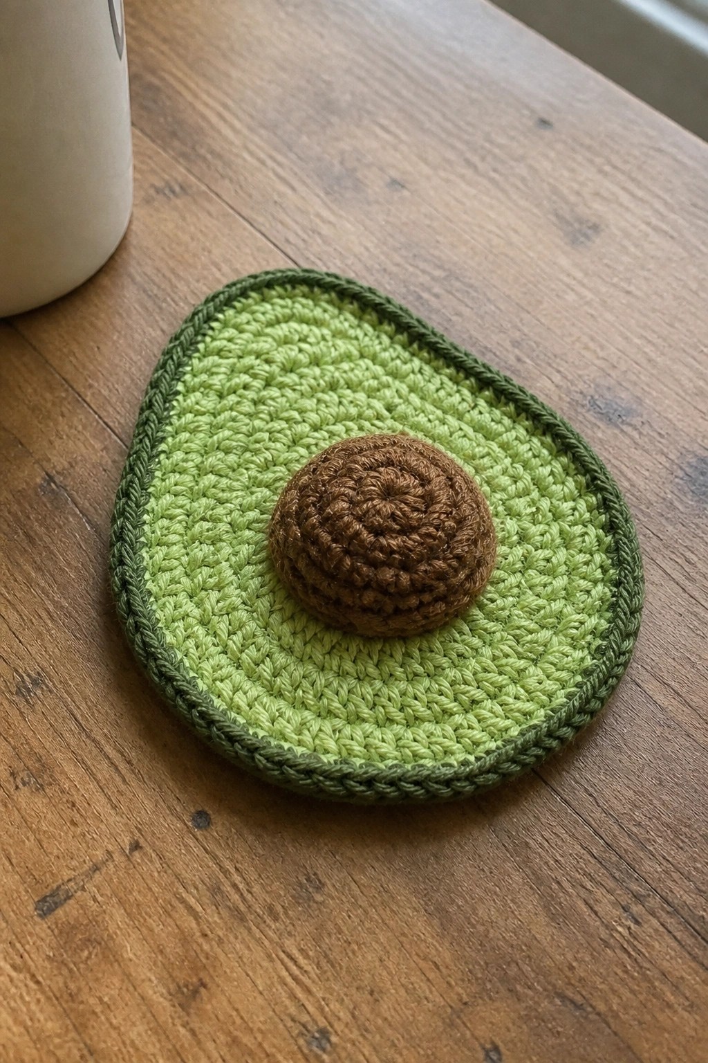 Crocheted green avocado coaster with brown center on wooden table near white mug