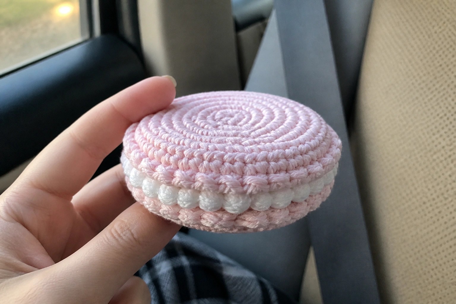 Hand holding pink crocheted macaron with white edge in car seat
