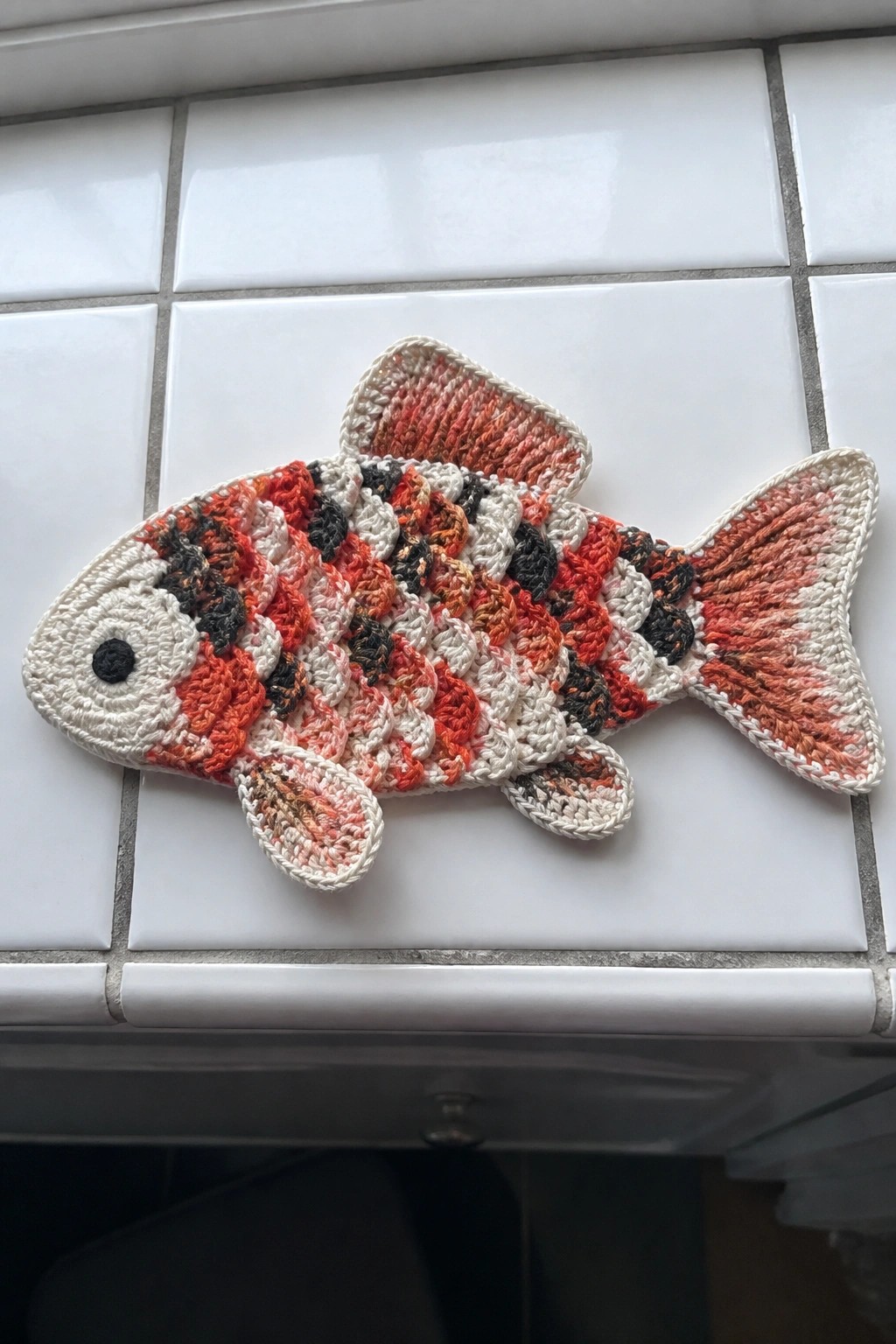 Colorful crocheted fish potholder with orange, red, black, and white yarn on tiles.