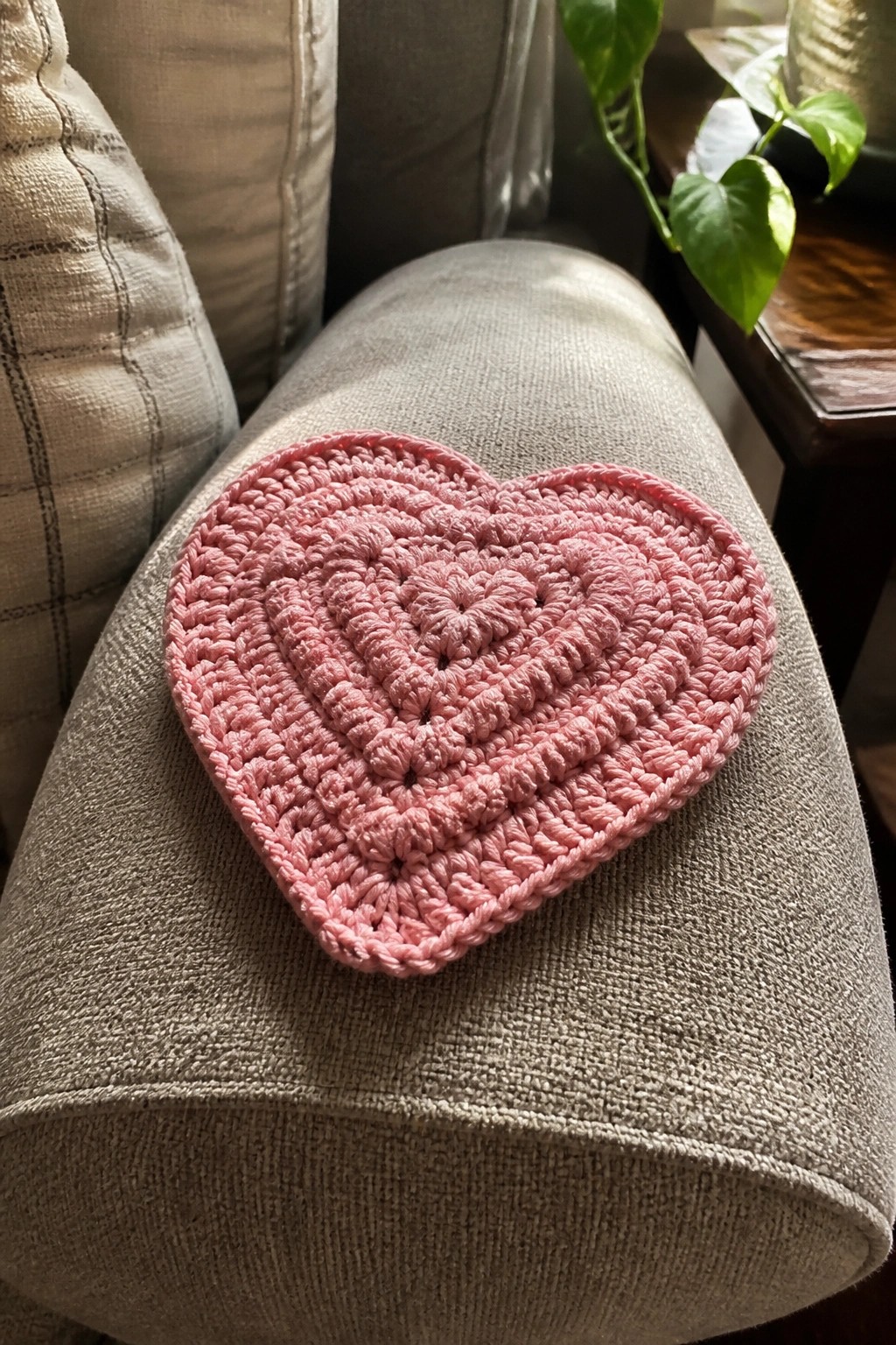 Pink crocheted heart resting on sunlit gray upholstered ottoman with plant nearby.