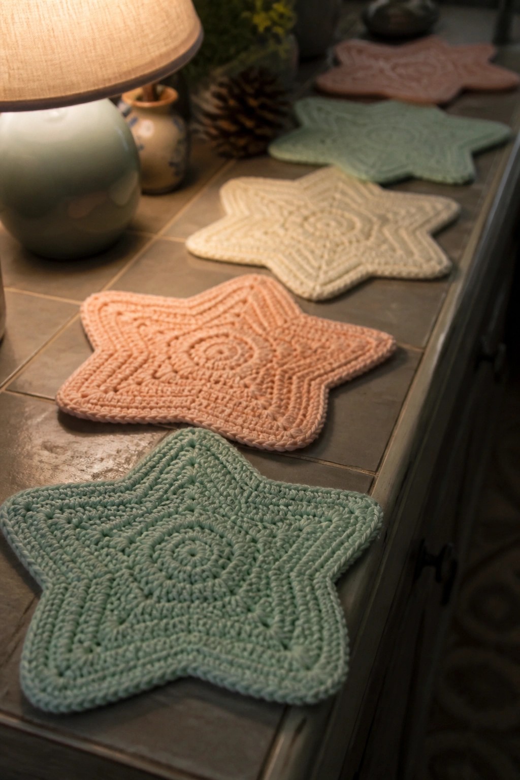 Pastel crocheted star coasters lined on gray tiles beside a lit lamp.