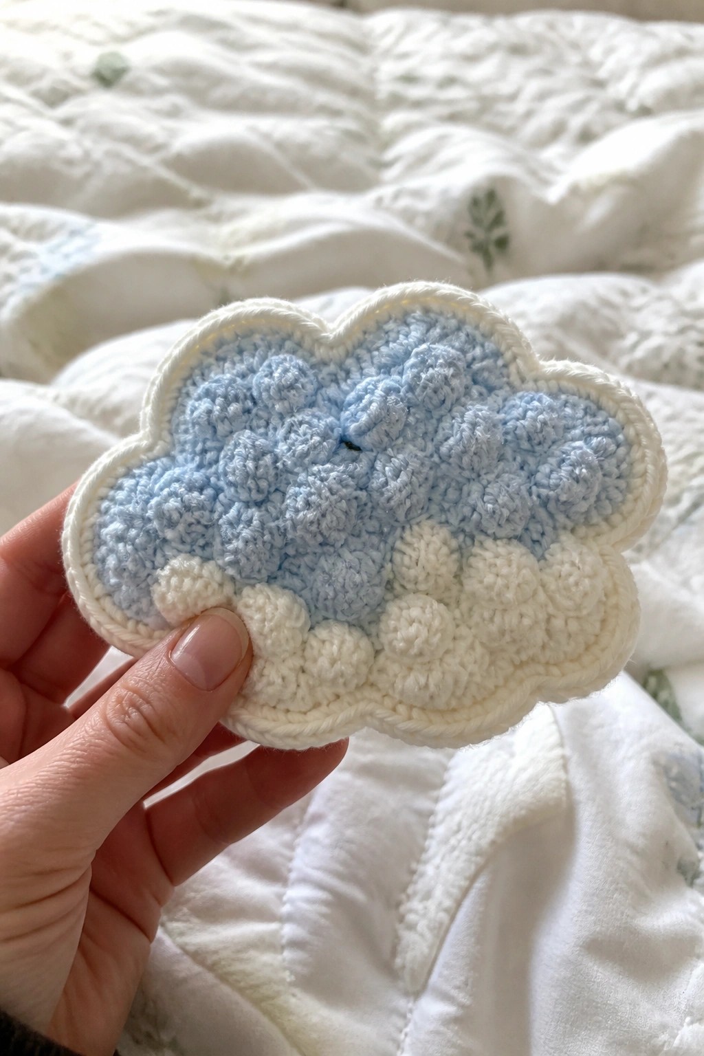Hand holding light blue and white bobble-stitch crocheted cloud over quilted bedding.