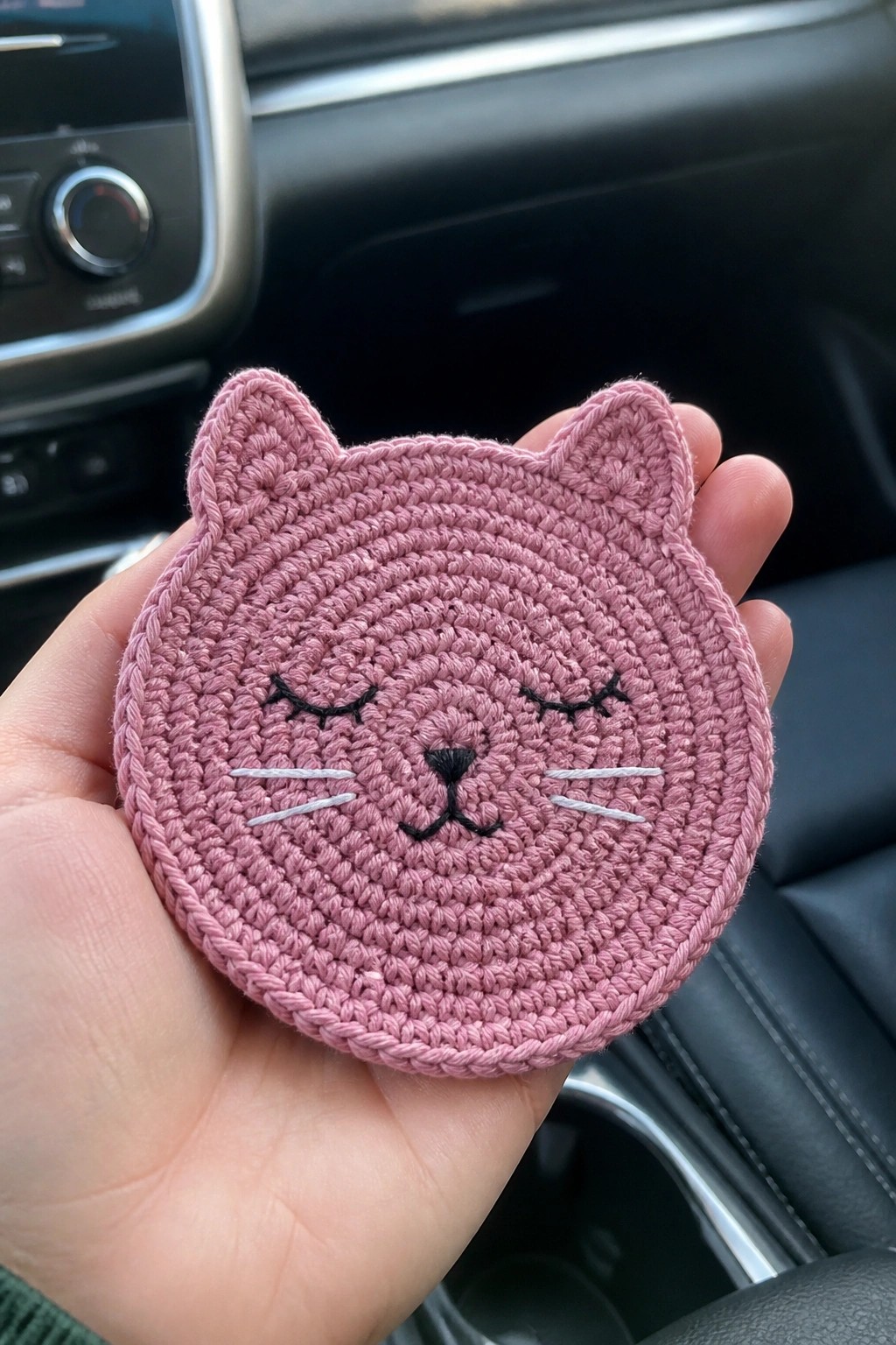 A hand holding a pink crocheted circular coaster with cat ears and an embroidered face showing closed eyes, whiskers, and a nose.