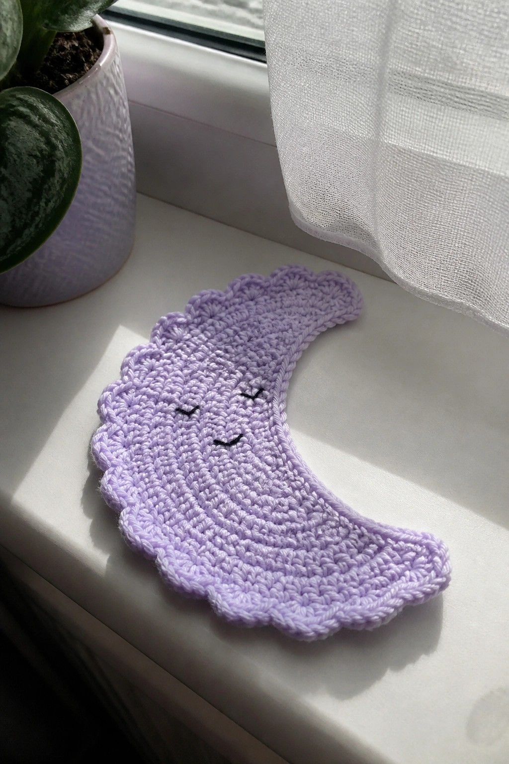 Purple crocheted crescent moon with embroidered face on sunny windowsill beside plant