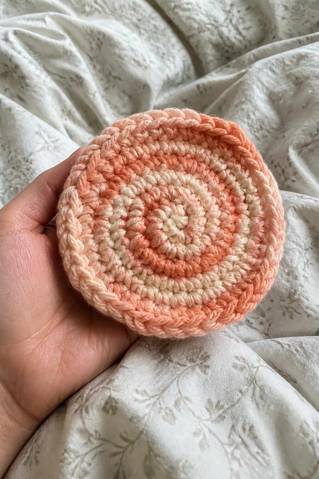 Hand holding a crocheted peach circular mat on white floral fabric.