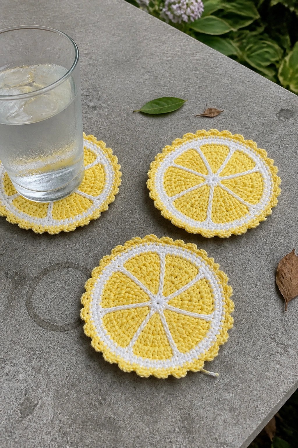 Three yellow crocheted lemon-slice coasters on gray stone table with water glass