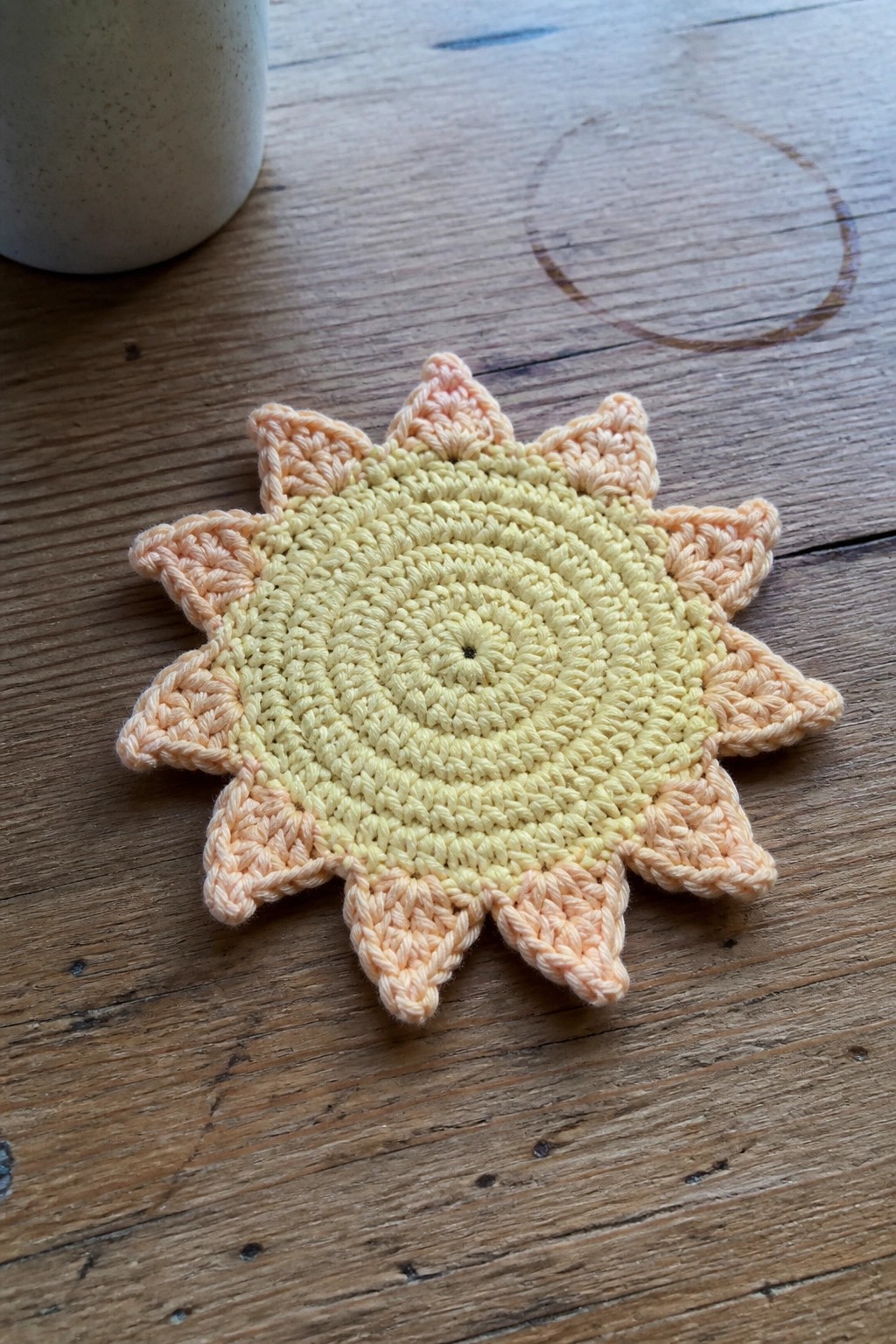A crocheted sun-shaped coaster in yellow and peach rests on a wooden table.