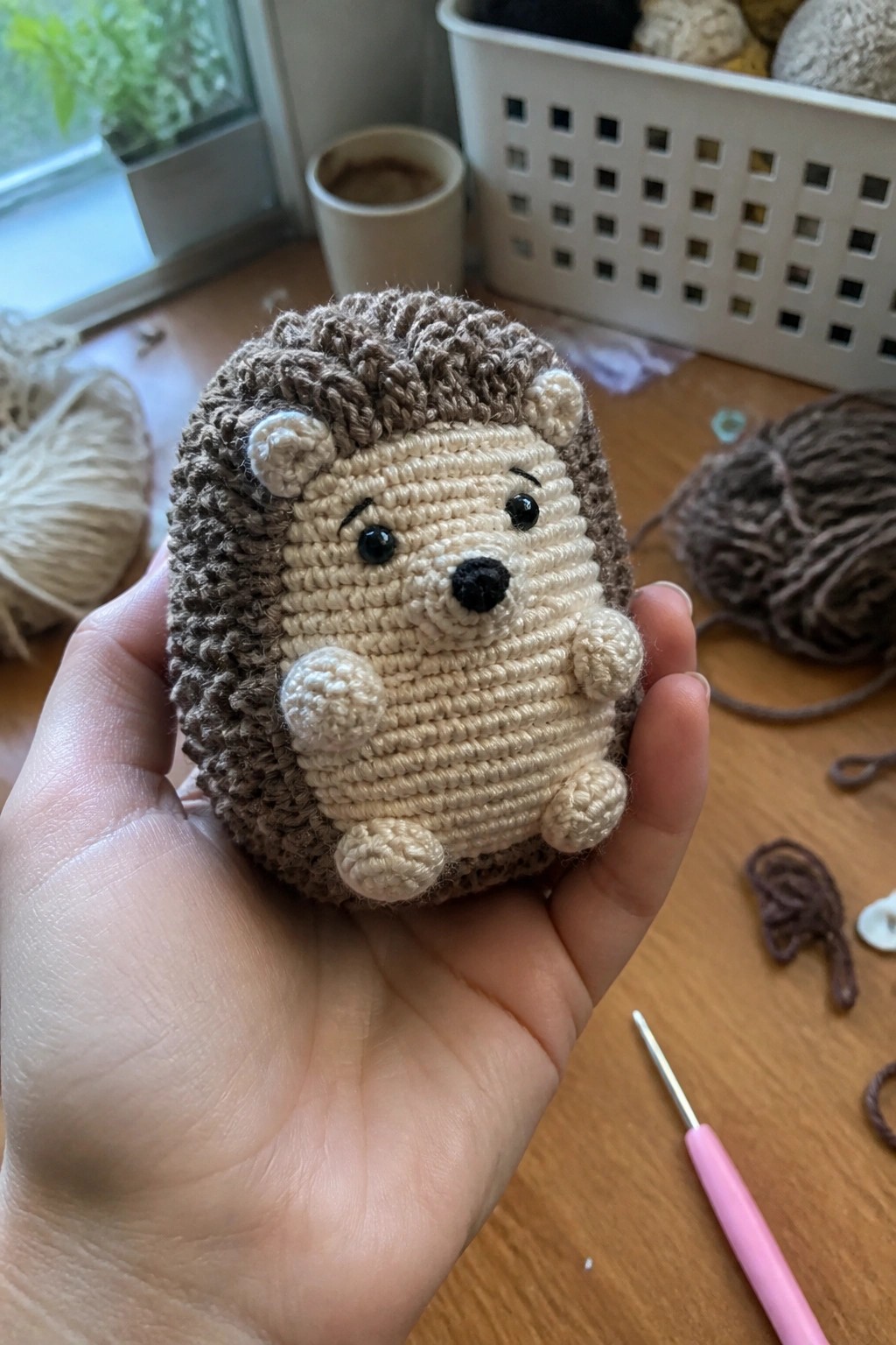 Hand holding crocheted beige-brown hedgehog amid yarn, hook, and supplies on wooden table
